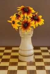 Yellow flowers of perennial rudbeckia in a wooden vase in the shape of a chess rook  on a chessboard on a brown background