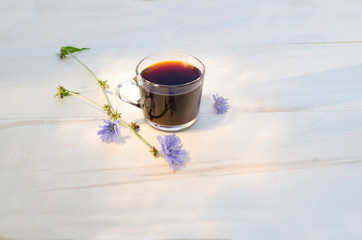 drink chicory in a transparent glass cup and blue flowers of the chicory plant on a white wooden background. A cup of chicory with highlights on the surface and reflection. Copy space.