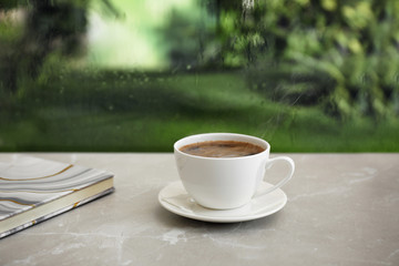 Cup of hot drink and notebook on stone windowsill against glass with rain drops, space for text