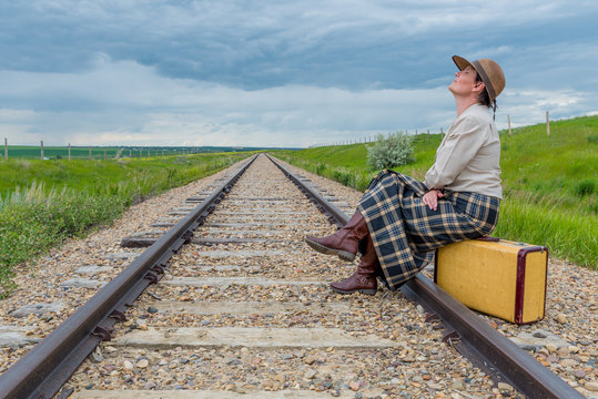 Young woman in historic dress sitting on vintage suitcase on railroad track with head back and eyes closed- journey concept