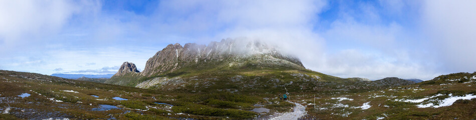 Fototapeta na wymiar A panorama of a Cradle Mountain and Tasmania's Overland Track with fog over the summit and a hiker on the trail with some snow and water on the ground
