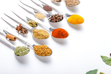 Various spices ( cumin, coriander, curry, paprika, chili, turmeric cinnamon, fenugreek, cardamom, basil leaf, parsley, cloves )  in  spoons on white background. Top view with copy space.