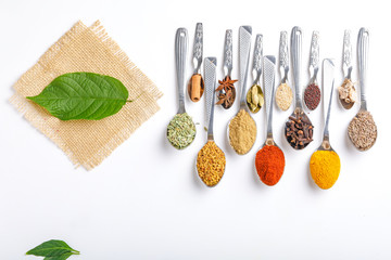 Various spices ( cumin, coriander, curry, paprika, chili, turmeric cinnamon, fenugreek, cardamom, basil leaf, parsley, cloves )  in  spoons on white background. Top view with copy space.