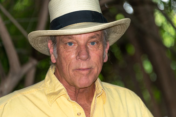 Portrait of good looking senior man in his 60s outdoors in backyard, wearing hat and yellow shirt.  Looking at camera