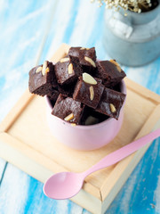 Piece of brownie almond in pink cup with pink spoon and dry flower on blue wood table and blue background.