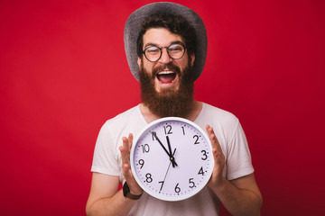 Photo of handsome excited bearded man, wearing a cowboy hat, holding clock  standing over red...