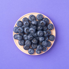 Ripe blueberries in the wooden bowl. Top view, summer berries on a purple background