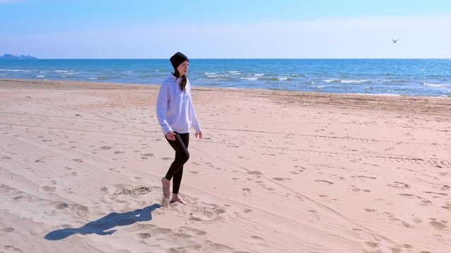 Young girl in hoodie, hat and leggins does yoga exercises on sea sand beach in warmy weather. She makes balance poses stratching her legs before training.