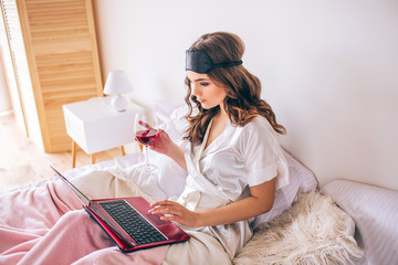 Young woman with dark hair sitting on bed in bedroom and working. Remote work in morning. Woman in gressing gown hold glass of red wine. Calm peaceful and concentrated.