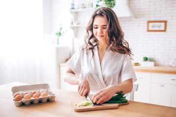 Young woman with dark hair standing in kitchen and cutting green onion. Eggs besides. Morning daylight. Alone in kitchen.