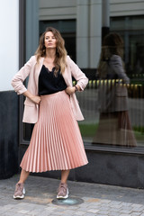 girl model in coral color clothes. Pleated long skirt, jacket, sneakers, black blouse. Long wavy hair. A very beautiful European woman stands on the street near a glass case.