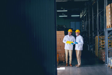 Two smiling warehouse workers in white uniforms and yellow helmets on heads standing and talking...
