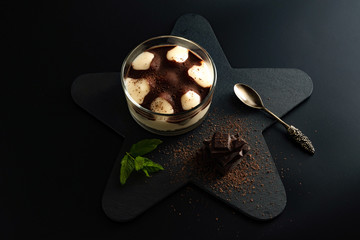 Cheesecake dessert in a jar on a black stone plate on the black background. Decorated with black chocolate pieces, grated chocolate and fresh mint.