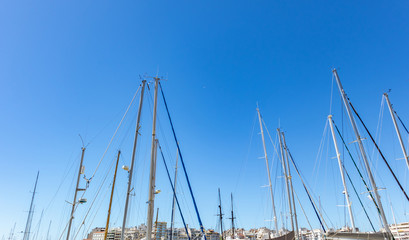Yachts mast tops against the clear blue sky at marina Zeas, Greece. Vertical lines with ropes, city background