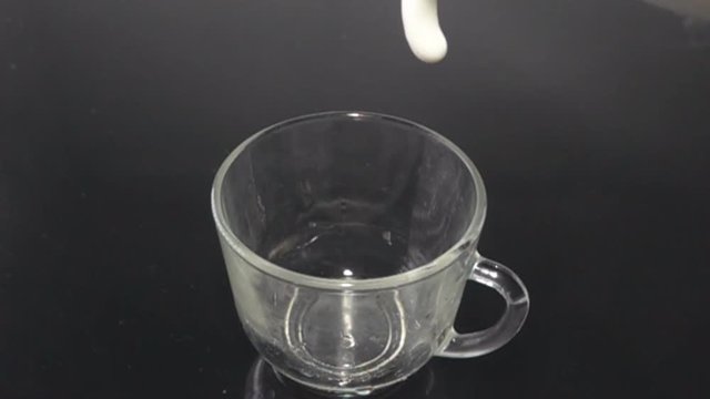 Butter milk (kefir, cultured yoghurt) flows into a transparent cup that stands on a black surface, dairy products, sour milk products, healthy diet. Super slow motion 1000 fps