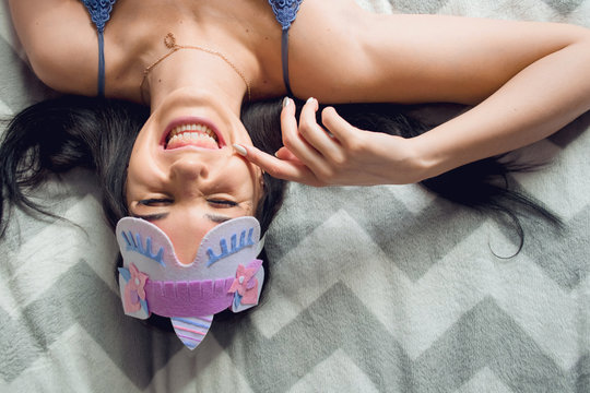 Young smilling brunette woman with long hair in blue nightdress and mask for sleeping with unicorn lying in bed with gray wave. Top view portrait of cheerful girl in eye mask and sleepwear. Copy space