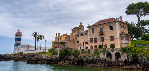 Obraz premium Beautiful traditional Portuguese building & a blue & white lighthouse on a rocky coast, taken in Cascais on a cloudy day.