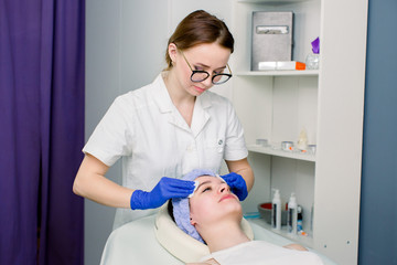 Obraz premium Portrait of professional woman cosmetologist wiping with cotton discs face of female client lying on white couch. Dermatologist in uniform in process of doing cosmetic procedure.