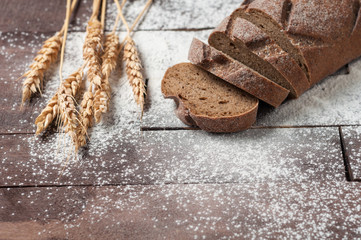 Sliced loaf of dark homemade bread dusted with flour near bunch of wheat is lying on old brown boards