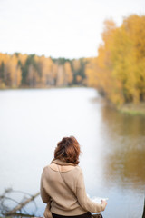 Fototapeta premium Girl sitting by the water on the lake, from the back, brunette, in beige clothes, Golden autumn.
