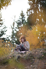 Fototapeta premium A beautiful brunette girl with curls, autumn in the Park by the water reading a book, clothes beige wool large knit, scarf warm, wind blowing, yellow foliage.