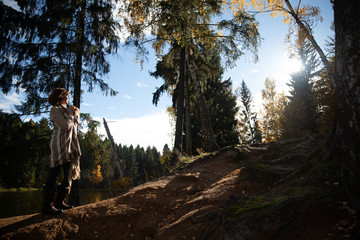 The girl climbs the mountain in the autumn forest near the lake towards the sun in brown clothes with a beige wool blanket.