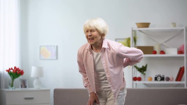 Old lady standing up from sofa suddenly feeling sharp lower back pain, spasm