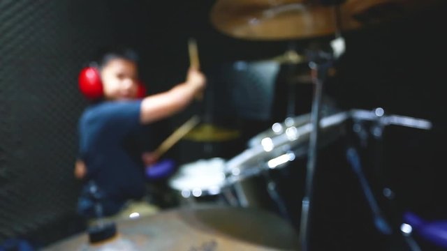 blurry image of Asian boy put blue tshirt and red headphone learning and play drum set with wooden drumsticks in music room.