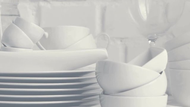 Stacked white porcelain dishes, plates, cups, saucers, mugs in restaurant kitchen interior. Slow pan. White brick wall background.	Selective focus.