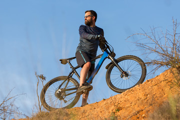 Cyclist in shorts and jersey on a modern carbon hardtail bike with an air suspension fork rides off-road on the orange-red hills at sunset evening in summer	