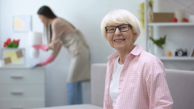 Smiling aged woman looking at camera, housekeeper wiping dust in room, cleaning