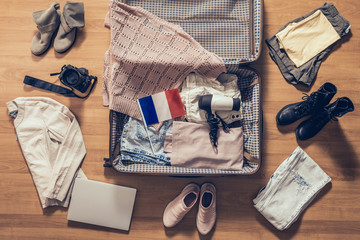 Woman's clothes, laptop, camera and flag of France lying on the parquet floor near and in the open suitcase. Travel concept