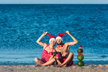 bearded man and girl in Santa clothes are sitting on sea beach with cups in their hands and snowman from watermelons. Summer Christmas Celebration.