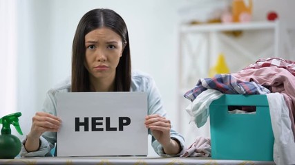 Upset woman showing help word on cardboard, laundry basket with clothes on table