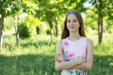 A young blonde girl in a pink t-shirt with arms crossed is standing against a background of green forest