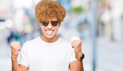 Young handsome man with afro hair wearing sunglasses celebrating surprised and amazed for success with arms raised and open eyes. Winner concept.