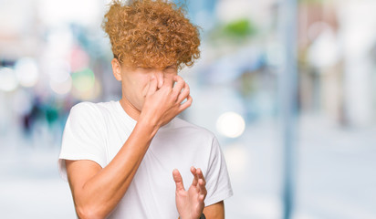 Young handsome man with afro hair wearing casual white t-shirt smelling something stinky and...