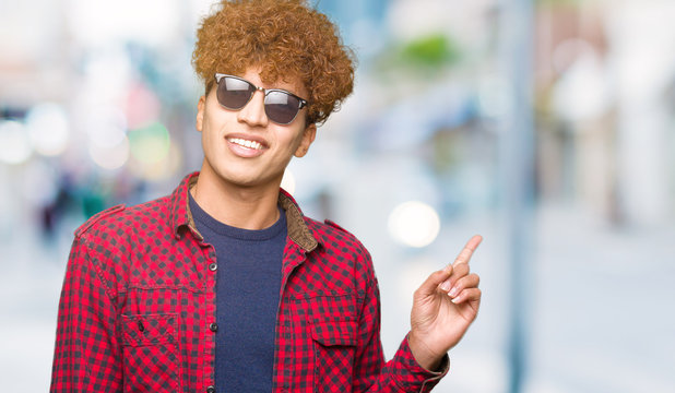 Young handsome man with afro hair wearing sunglasses with a big smile on face, pointing with hand and finger to the side looking at the camera.