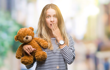 Young beautiful blonde woman holding teddy bear plush over isolated background cover mouth with...