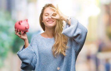 Young beautiful blonde woman holding piggy bank over isolated background with happy face smiling...