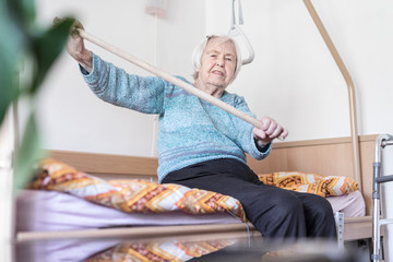 Elderly 96 years old woman exercising with a stick sitting on her bad. Geriatric health care home...