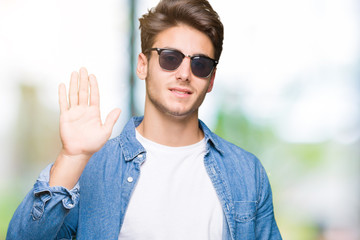 Young handsome man wearing sunglasses over isolated background Waiving saying hello happy and smiling, friendly welcome gesture