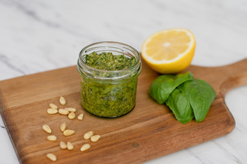 Homemade pesto in a small jar on a wood cutting board; white marble countertop; lemon, basil, and pine nuts scattered around jar.