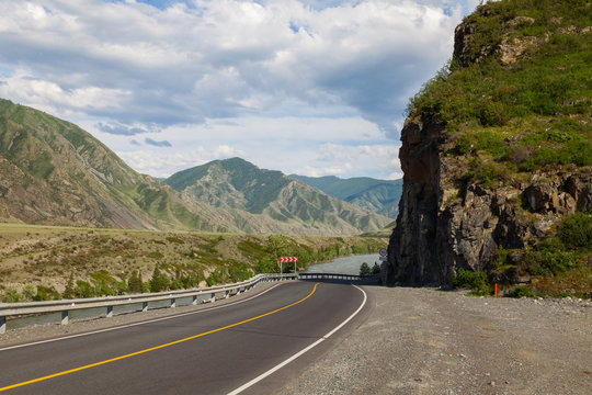 A new winding asphalt black road with a yellow dividing strip between the rocks and the Katun River in the Altai mountains under a clear blue sky with clouds and a field covered with green grass