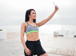 Young fit brunette woman in sport wear listening music with headphones and makes selfie with smartphone in her hands on  beach at sunrise