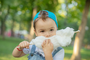 Adorable little kid eating candy-floss outdoors at summer park. Boy holding cotton candy. Little boy eating cotton candy on a stick. Happy childhood. Funny kid in a park. Kids love sweets