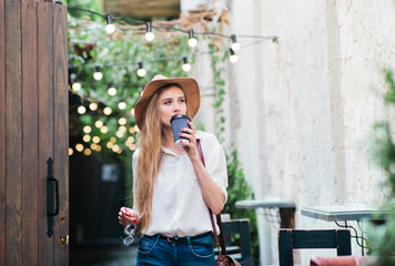 Young attractive woman in casual style clothes walking along the street of the old city with a cup of coffee on the go