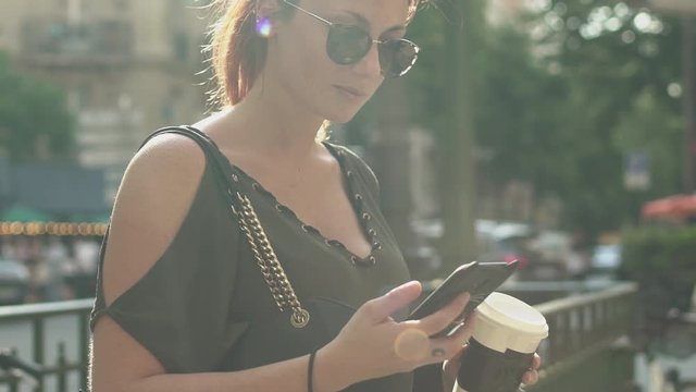 Young attractive woman writing a text message on her smartphone at the subway exit in street, holding her coffee in other hand, during sunny summer afternoon in Paris. Trendy and cool, tilt up.