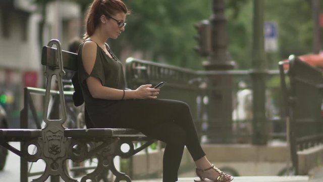 Attractive caucasian impatient woman with glasses, freckles, piercings and red hair waiting and watching at her smartphone sitting on street bench, during sunny summer in Paris. Slow motion.