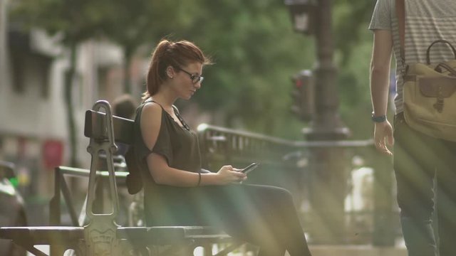 Attractive caucasian impatient woman with glasses, freckles, piercings and red hair waiting and watching at her smartphone sitting on street bench, during sunny summer in Paris. People passing by.